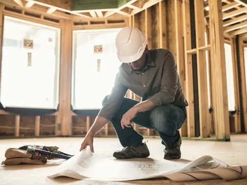 A contractor kneeling over rolled framing plans inside a house under construction
